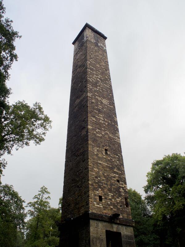 Looking up towards a yellowish, square, brick chimney set against a grey sky.